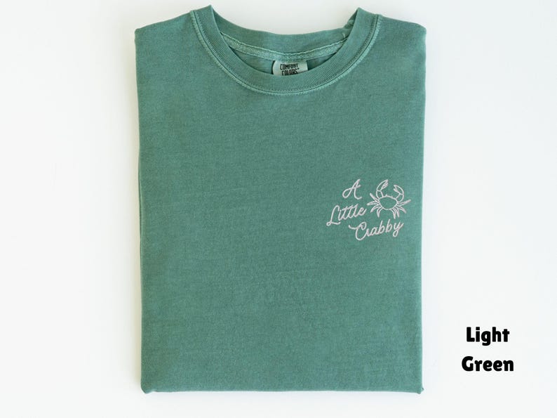 May include: Light green t-shirt with a crew neck. The shirt has the embroidered phrase "A Little Crabby" with a crab graphic on the front. The shirt is folded neatly on a white surface.