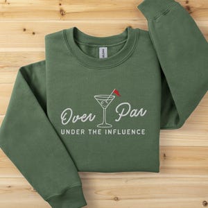 May include: Green sweatshirt with the embroidered text "Over Par Under the Influence" and a martini glass with a golf flag. The long-sleeved crew neck top is laid on a wooden surface.