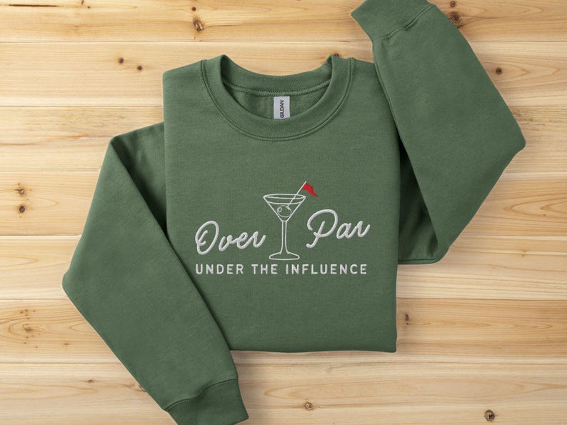 May include: Green sweatshirt with the embroidered text "Over Par Under the Influence" and a martini glass with a golf flag. The long-sleeved crew neck top is laid on a wooden surface.