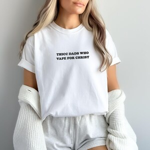 Puede incluir: Camiseta blanca con el texto negro "THICC DADS WHO VAPE FOR CHRIST". La camiseta se usa con pantalones cortos grises y una rebeca blanca. Las mangas est&aacute;n enrolladas.