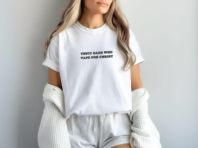Puede incluir: Camiseta blanca con el texto negro "THICC DADS WHO VAPE FOR CHRIST". La camiseta se usa con pantalones cortos grises y una rebeca blanca. Las mangas est&aacute;n enrolladas.