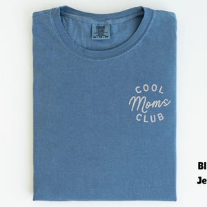 May include: A folded blue jean colored t-shirt with the words "COOL Moms CLUB" in white script on the front. The shirt is made of a soft material and has a crew neck.