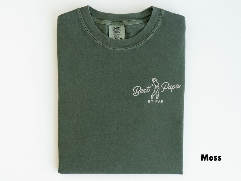 Op de afbeelding: Mosgroen T-shirt met de tekst "Best Papa" en een golf-thema afbeelding. Het shirt is netjes opgevouwen en heeft een label bij de halslijn. De tekst "BY PAR" staat onder de afbeelding. Het shirt is gemaakt van zacht materiaal.