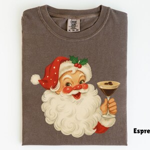 May include: A folded, dark gray t-shirt featuring a vintage illustration of Santa Claus holding a martini glass with coffee beans. Santa wears a red hat with white polka dots. The word "Espresso" is printed on the shirt.