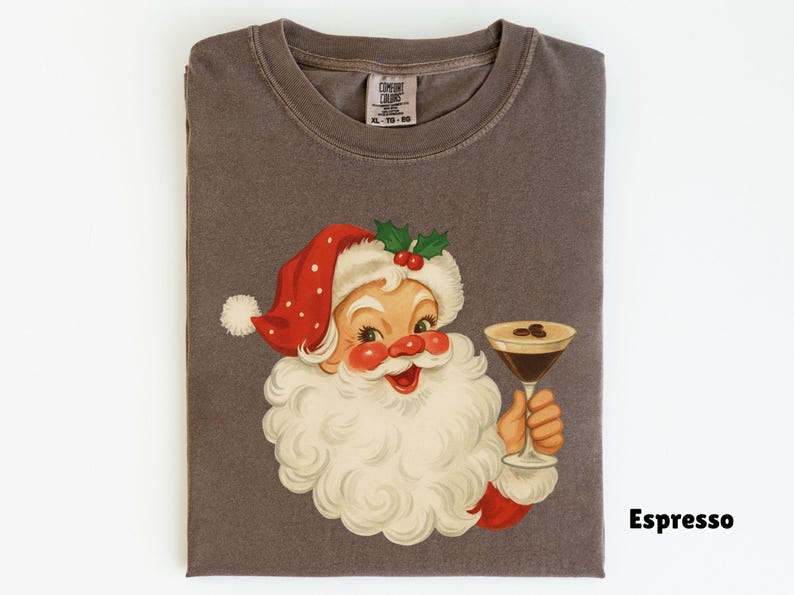May include: A folded, dark gray t-shirt featuring a vintage illustration of Santa Claus holding a martini glass with coffee beans. Santa wears a red hat with white polka dots. The word "Espresso" is printed on the shirt.