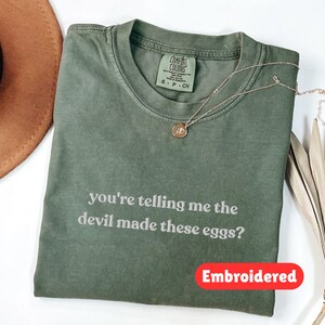 May include: Olive green t-shirt with the phrase "you're telling me the devil made these eggs?" embroidered in white. A gold necklace with a pendant rests on the shirt. A brown hat and dried leaves are also in the image.