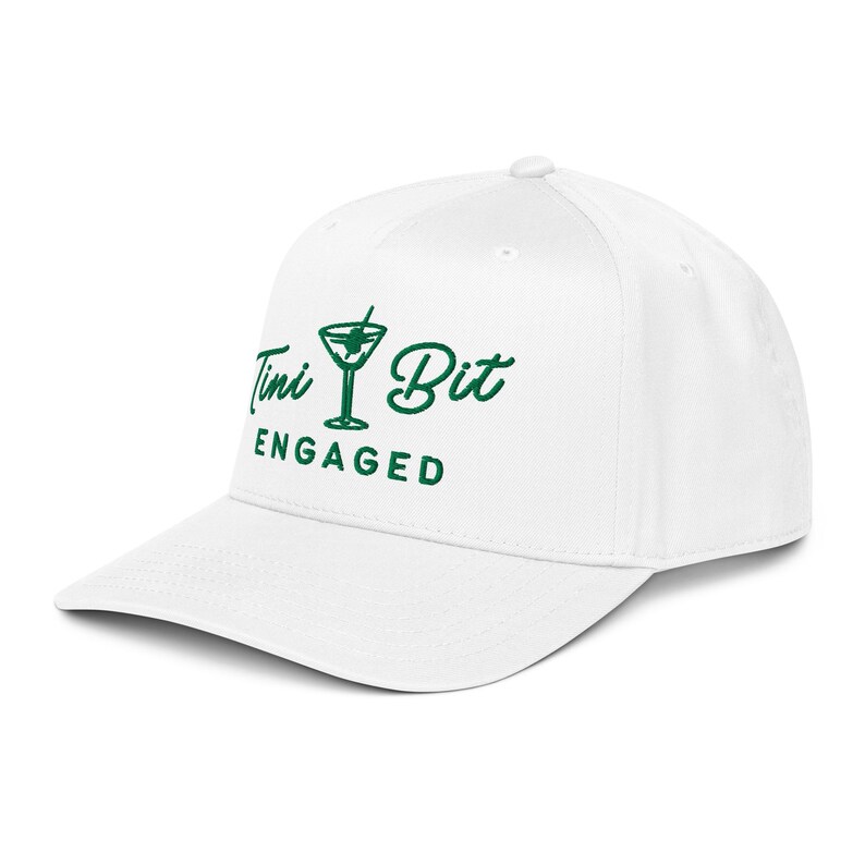 Puede incluir: Gorra de b&eacute;isbol blanca con visera curva. La gorra presenta el texto "Tini Bit Engaged" en verde, junto con un gr&aacute;fico de copa de martini verde. Un accesorio casual y cl&aacute;sico.