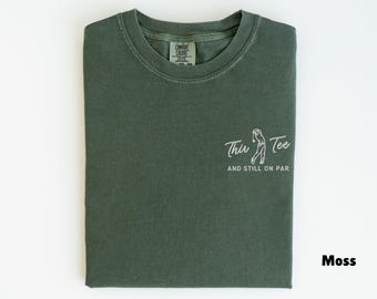 May include: Moss green t-shirt with the text "Thin Tee and still on par" and a graphic of a golfer. The shirt is folded neatly on a white surface. The shirt is a Comfort Colors brand.