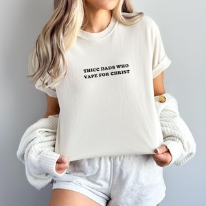 Puede incluir: Camiseta color crema con el texto negro "THICC DADS WHO VAPE FOR CHRIST" centrado en la parte delantera. La camiseta tiene las mangas enrolladas y se combina con pantalones cortos grises. Un su&eacute;ter blanco est&aacute; sobre los hombros.