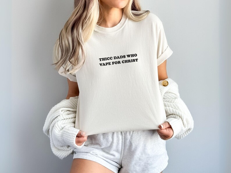 Puede incluir: Camiseta color crema con el texto negro "THICC DADS WHO VAPE FOR CHRIST" centrado en la parte delantera. La camiseta tiene las mangas enrolladas y se combina con pantalones cortos grises. Un su&eacute;ter blanco est&aacute; sobre los hombros.