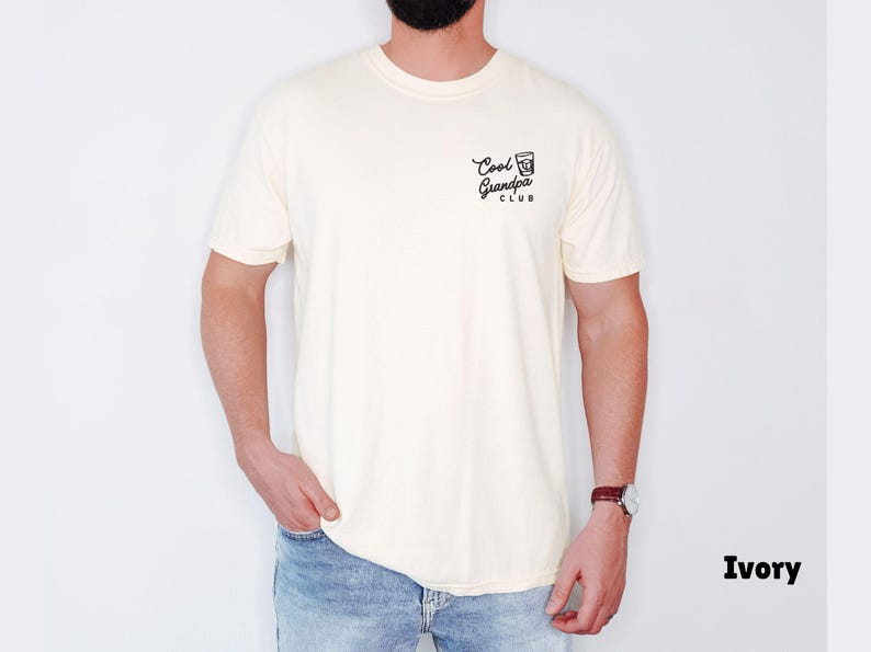 May include: Ivory-colored t-shirt with the text "Cool Grandpa Club" in black script and a small graphic. The shirt is a crew neck and is being worn with blue jeans. A watch with a brown leather band is visible.