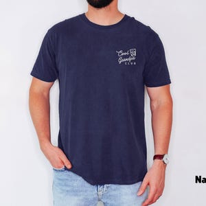 May include: Navy blue t-shirt with the text "Cool Grandpa Club" printed on the front. The shirt has a classic crew neck and short sleeves. The image shows a person wearing the shirt with light blue jeans.