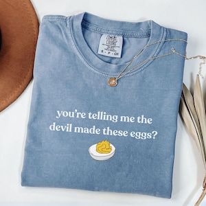 May include: A light blue t-shirt with the text "you're telling me the devil made these eggs?" and an illustration of a deviled egg. A gold necklace and a brown hat are also in the image.