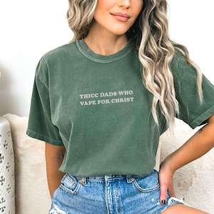 Puede incluir: Camiseta verde de manga corta con el texto "THICC DADS WHO VAPE FOR CHRIST" en gris claro. La camiseta est&aacute; hecha de un material suave y tiene un corte relajado.