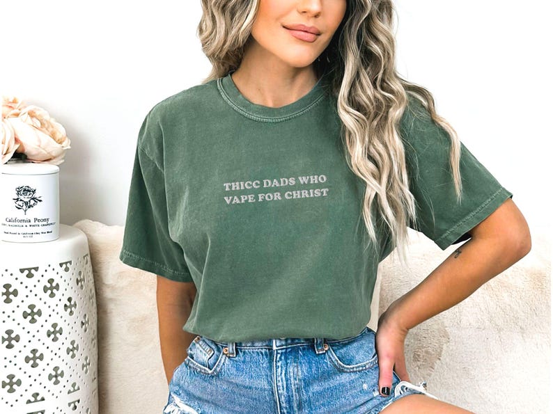 Puede incluir: Camiseta verde de manga corta con el texto "THICC DADS WHO VAPE FOR CHRIST" en gris claro. La camiseta est&aacute; hecha de un material suave y tiene un corte relajado.