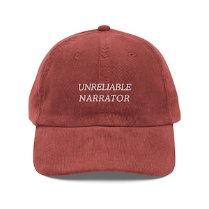 May include: A brick-red corduroy baseball cap with a curved brim. The text "UNRELIABLE NARRATOR" is embroidered in white on the front of the cap. The cap has a rounded crown and an adjustable strap.