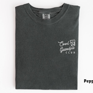 May include: Pepper-colored t-shirt with the text "Cool Grandpa Club" in white script and a graphic of a cocktail. The shirt is folded neatly on a white surface. The shirt is made of a soft, comfortable material.