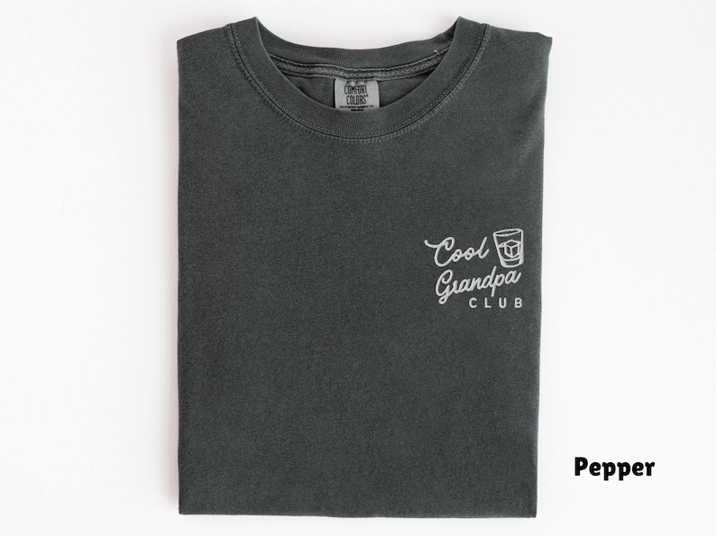 May include: Pepper-colored t-shirt with the text "Cool Grandpa Club" in white script and a graphic of a cocktail. The shirt is folded neatly on a white surface. The shirt is made of a soft, comfortable material.