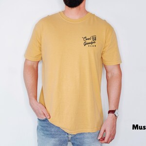 May include: Mustard-colored t-shirt with the text "Cool Grandpa Club" printed on the upper left chest. The shirt is a short-sleeved crew neck style. The model is wearing blue jeans and a watch.