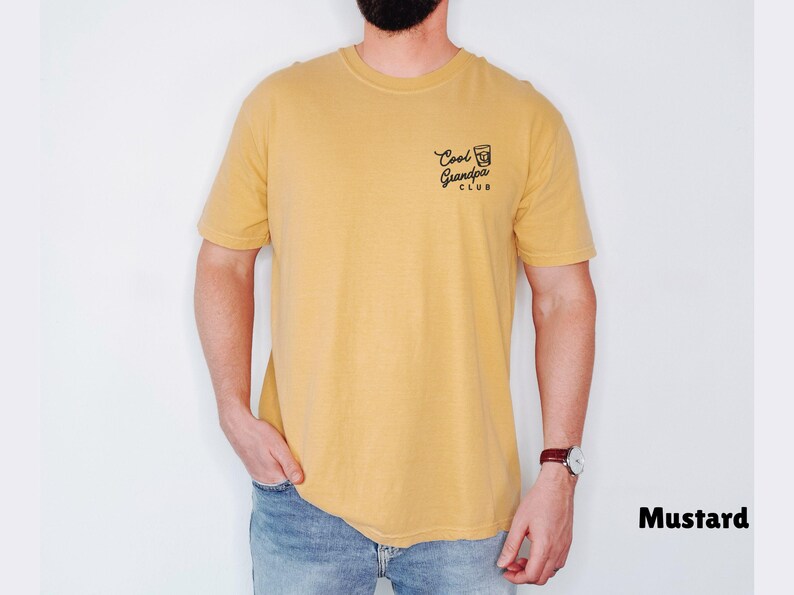 May include: Mustard-colored t-shirt with the text "Cool Grandpa Club" printed on the upper left chest. The shirt is a short-sleeved crew neck style. The model is wearing blue jeans and a watch.