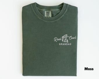 May include: Moss green t-shirt with the words "Reel Cool Grandad" in white script and a fish graphic. The shirt is folded neatly on a white surface. The Comfort Colors tag is visible.