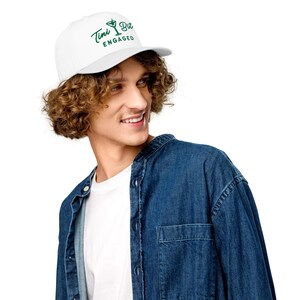 Puede incluir: Gorra de camionero blanca con visera curva. La gorra presenta el texto "Tini Bit ENGAGED" en verde, junto con un gr&aacute;fico de copa de Martini. Un accesorio casual.
