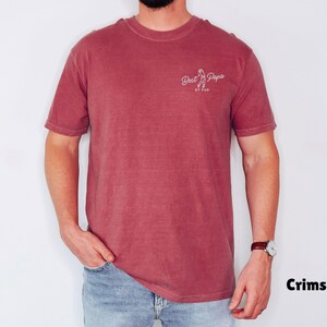 Op de afbeelding: Karmizijnrood T-shirt met de tekst "Best Papa" in witte letters. Het shirt heeft een ronde hals en is effen van kleur. Het model draagt een blauwe spijkerbroek en een horloge.