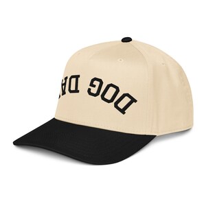 May include: A beige baseball cap with a black brim and the words "DOG DAD" embroidered in black. The cap has a curved brim and a small black button on top.