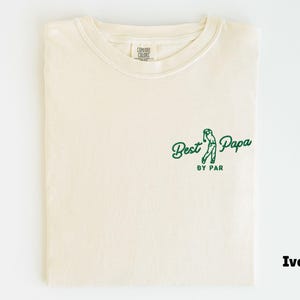 Op de afbeelding: Ivoor kleurig T-shirt met een ronde hals. Het shirt heeft een groene afbeelding van een golfer en de tekst "Best Papa by Par". Het shirt is netjes opgevouwen.