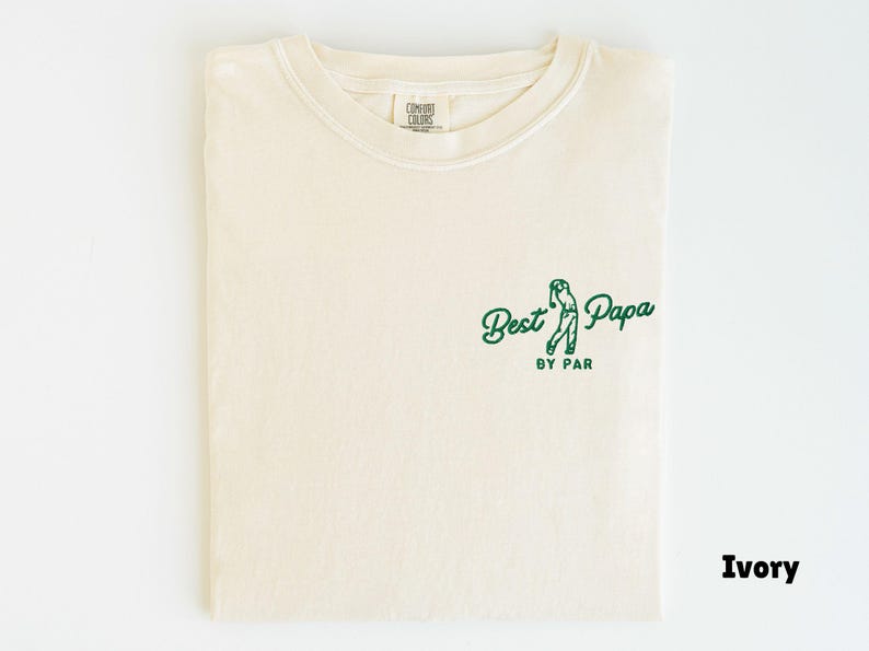 Op de afbeelding: Ivoor kleurig T-shirt met een ronde hals. Het shirt heeft een groene afbeelding van een golfer en de tekst "Best Papa by Par". Het shirt is netjes opgevouwen.