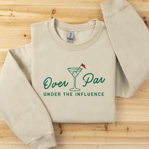 May include: Beige crewneck sweatshirt with the text "Over Par Under the Influence" in green, along with a martini glass graphic with a golf flag. The sweatshirt is folded on a wooden surface.
