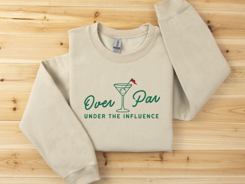May include: Beige crewneck sweatshirt with the text "Over Par Under the Influence" in green, along with a martini glass graphic with a golf flag. The sweatshirt is folded on a wooden surface.