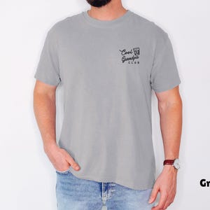 May include: A grey t-shirt with the text "Cool Grandpa Club" printed in black on the upper left chest. The shirt is short-sleeved and has a relaxed fit. The model is wearing blue jeans and a watch.