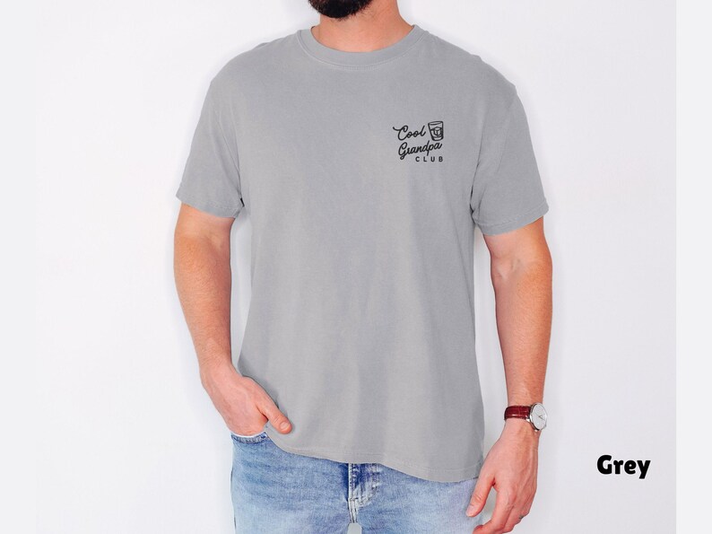 May include: A grey t-shirt with the text "Cool Grandpa Club" printed in black on the upper left chest. The shirt is short-sleeved and has a relaxed fit. The model is wearing blue jeans and a watch.