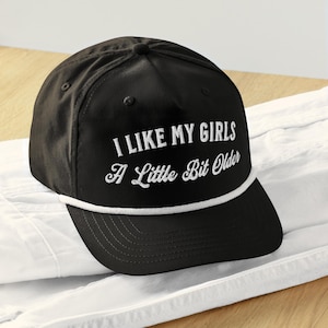 May include: Black baseball cap with a white rope trim and white embroidered text that reads "I like my girls a little bit older."