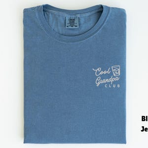 May include: Blue jean t-shirt with the text "Cool Grandpa Club" in white script and a graphic of a cocktail. The shirt is folded and has a tag in the collar.