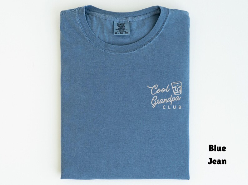 May include: Blue jean t-shirt with the text "Cool Grandpa Club" in white script and a graphic of a cocktail. The shirt is folded and has a tag in the collar.