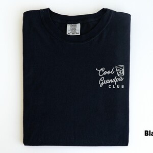 May include: Black t-shirt with the text "Cool Grandpa Club" in white script and a graphic of a whiskey glass. The shirt is folded and has a tag at the neckline. The word "Black" is printed in the lower right corner.
