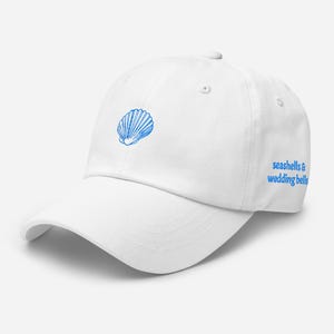 May include: White baseball cap with a blue seashell graphic on the front left and the text "seashells & wedding bells" on the right side. The cap has a curved brim and a button on top.
