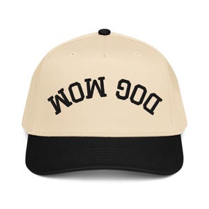 May include: A beige baseball cap with a black brim and a black button on top. The cap features the embroidered text "DOG MOM" in black, arched across the front. This hat is a stylish accessory for dog lovers.