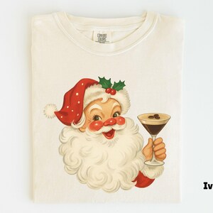 May include: Ivory-colored t-shirt featuring a vintage illustration of Santa Claus holding a cocktail. Santa is depicted with a red hat with white polka dots, a white beard, and rosy cheeks. The cocktail has coffee beans.