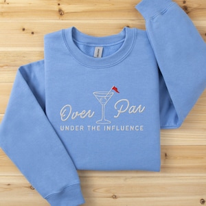 May include: A light blue sweatshirt with the embroidered phrase "Over Par Under the Influence" and a martini glass with a red flag. The sweatshirt is made of a soft material and is laid on a wooden surface.