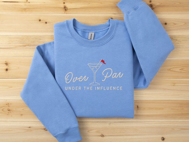 May include: A light blue sweatshirt with the embroidered phrase "Over Par Under the Influence" and a martini glass with a red flag. The sweatshirt is made of a soft material and is laid on a wooden surface.