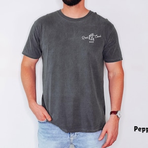Pu&ograve; includere: T-shirt color pepe con la scritta "Reel Cool Dad" in corsivo bianco. La maglietta &egrave; un girocollo a maniche corte. Il modello indossa jeans blu e un orologio.