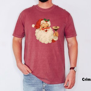 May include: Crimson-colored t-shirt featuring a vintage illustration of Santa Claus holding a cocktail. Santa is depicted with a white beard, red hat, and a jolly expression. The shirt is a solid color with the word "Crimson" printed in black.