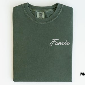 May include: A folded moss green t-shirt with the word "Funcle" embroidered in white script. The shirt has a crew neck and a tag visible at the back of the neck. The word "Moss" is printed in the bottom right corner.