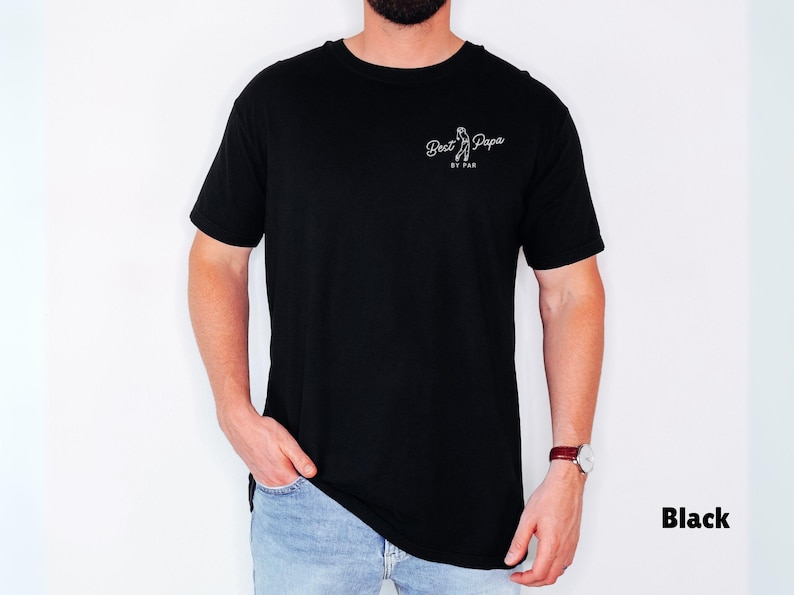 Op de afbeelding: Zwart T-shirt met de tekst "Best Papa" en een witte golfclubafbeelding. Het shirt is gemaakt van een zacht materiaal en is geschikt voor vrijetijdskleding. De tekst is wit en het shirt wordt door een persoon gedragen.
