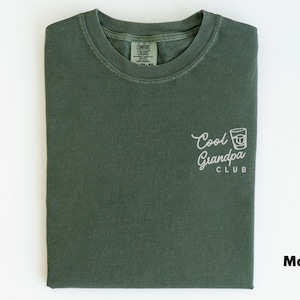May include: Moss green t-shirt with the words "Cool Grandpa Club" and a graphic of a drink in a glass. The shirt is folded and has a tag in the upper center. The word "Moss" is in the lower right corner.