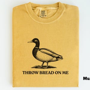 Puede incluir: Camiseta de color mostaza con un gráfico de un pato y el texto "THROW BREAD ON ME". El pato es negro y marrón, de pie sobre hierba. La camiseta está doblada y tiene cuello redondo.