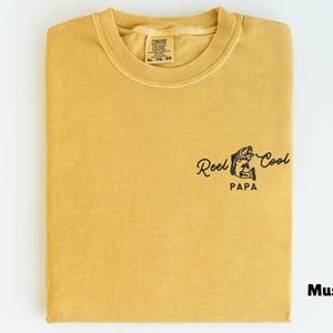 Op de afbeelding: Mosterdkleurig T-shirt met de tekst "Reel Cool Papa" en een visafbeelding. Het gevouwen shirt heeft een label met de tekst "Comfort Colors XL 10-20".